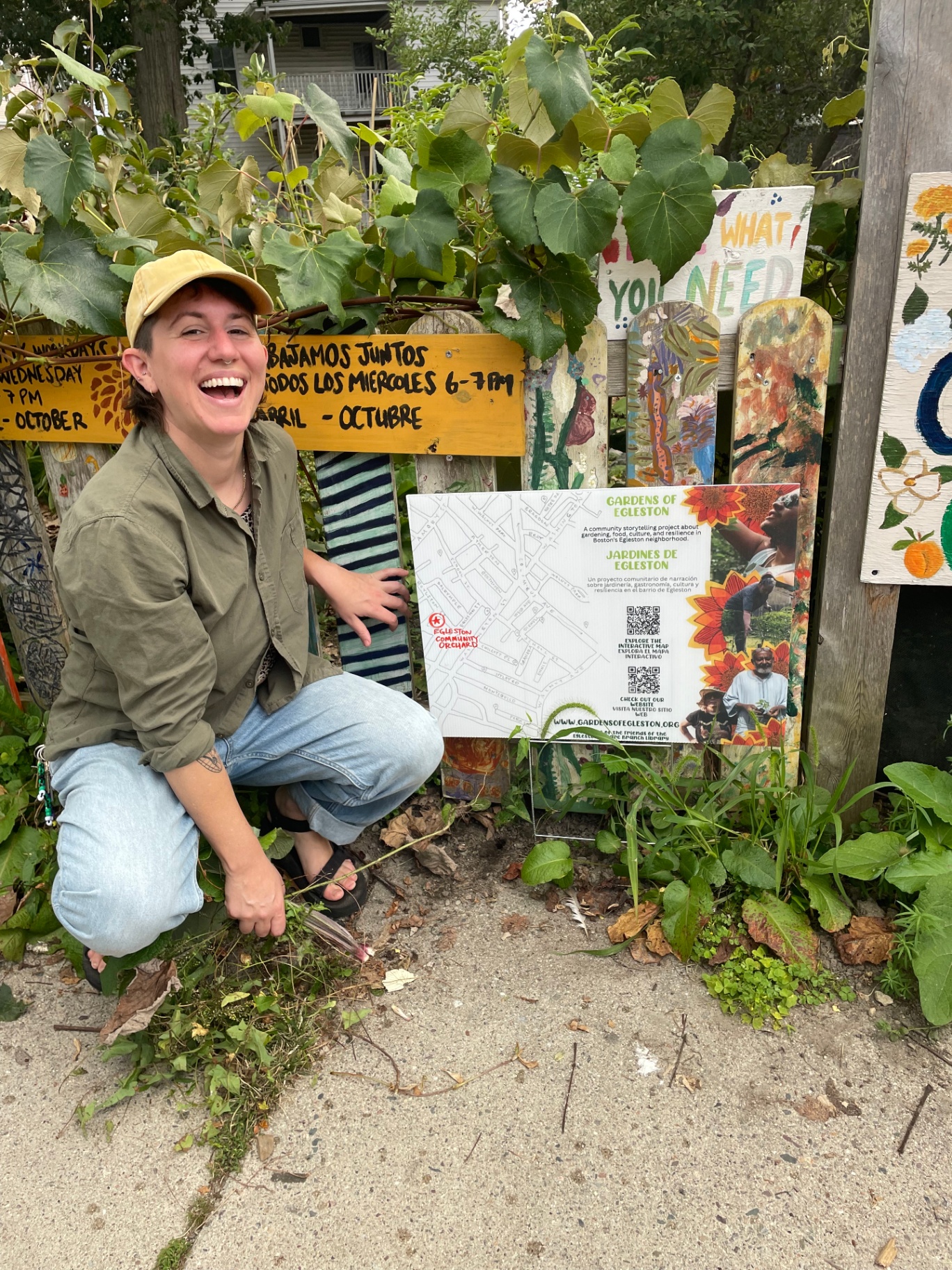A smiling person in a yellow cap and olive shirt crouched beside a hand-painted Gardens of Egleston / Jardines de Egleston signboard at the Egleston Community Orchard, surrounded by leafy vines and bilingual painted fence boards.